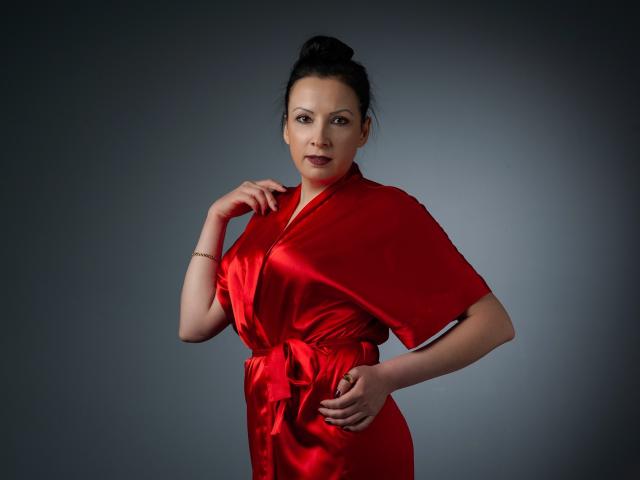 ElizabethSo wearing a shiny red satin robe, hand on hip, other hand touching her chin, dark hair in a bun, subtle smile, gray background, soft lighting.