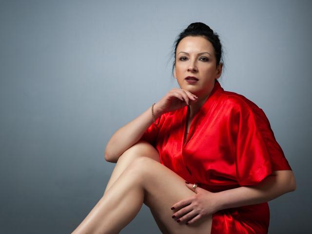 ElizabethSo, in a shiny red silk robe, sits with one knee up, hand on chin, looking at the camera. She has pale skin, dark hair in a bun, and is sexy.