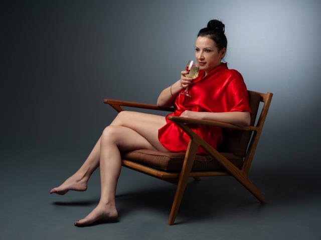 Elizabeth: Então, nua, usando um roupão de seda vermelho, sentada em uma cadeira de madeira, segurando uma taça de champanhe, pernas cruzadas, cabelo escuro preso em um coque, fundo cinza, foto de estúdio, pés descalços, sexy..