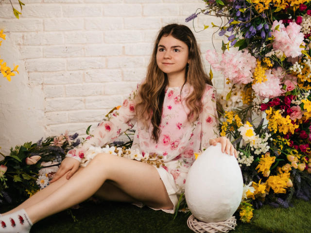 TinaRedly with long brown hair, pink floral dress, white socks, sitting on green grass, holding white egg-shaped object, surrounded by colorful flowers, white brick wall background.