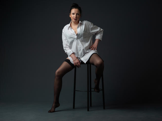 ElizabethSo sits on a stool, wearing an open white shirt, black garter stockings, and no pants, exposing her pussy. Dark hair in a bun, gray background.