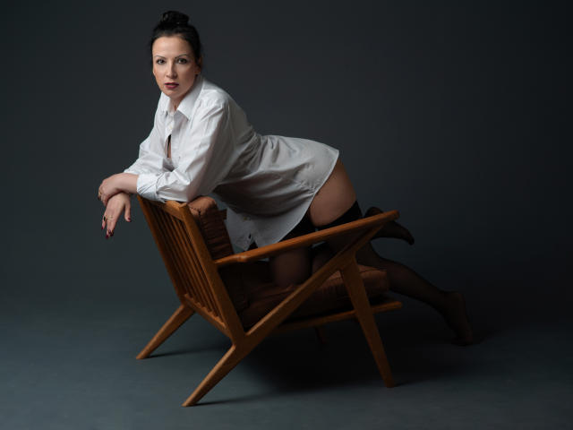 ElizabethSo with dark hair, wearing a white shirt and black stockings, leans on a wooden chair, exposing her bare ass and pussy. Dark background, studio lighting.