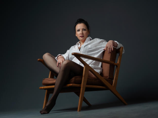 ElizabethSo, sitting in a wooden chair, wearing a white shirt, black tights, and no pants, showing her ass and crossed legs, dark hair in a bun, looking confident.