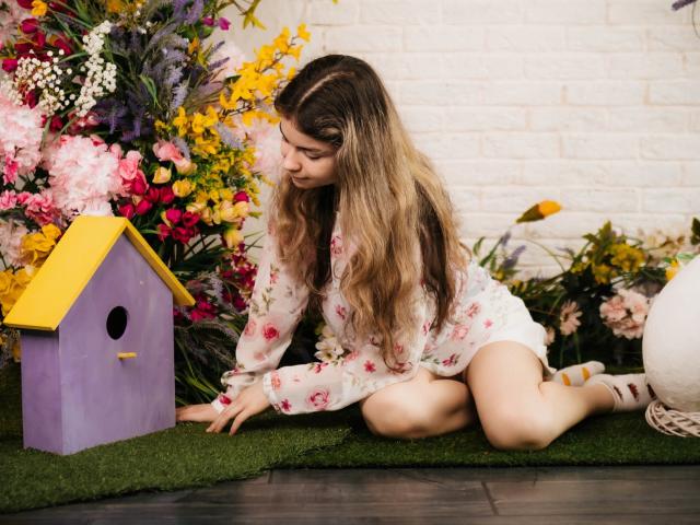 TinaRedly with long wavy blonde hair, floral shirt, and white shorts, kneeling on green grass, touching a purple birdhouse with a yellow roof, surrounded by colorful flowers.