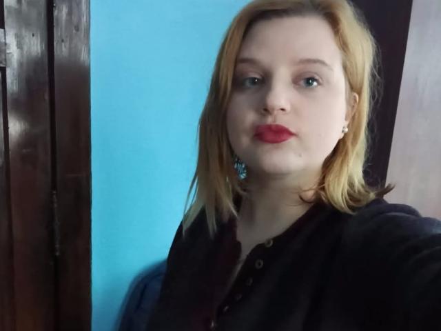 HospotsForU has pale skin, blonde hair, and red lipstick. She's wearing a black shirt, blue earrings, and a buttoned collar, with a hand near her face.
