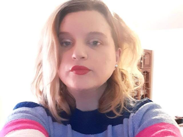 HospotsForU has blonde hair, red lipstick, and is wearing a blue and pink striped sweater with small earrings. Her face is slightly illuminated, with a soft, relaxed expression.
