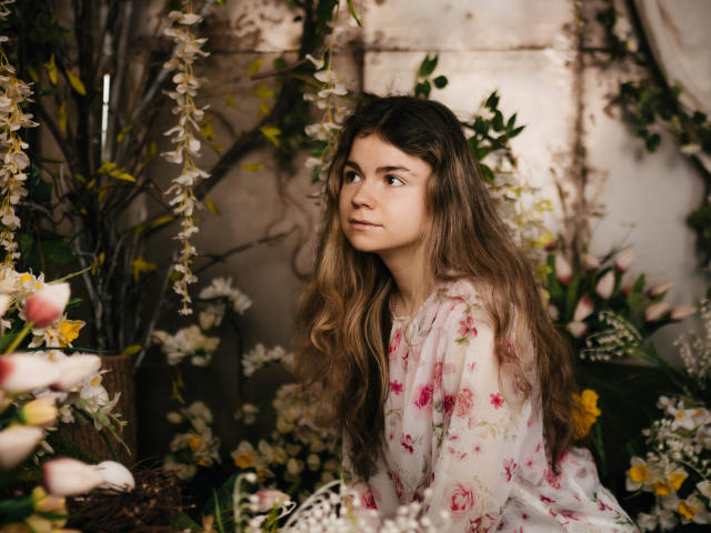 TinaRedly with long brown wavy hair, wearing a floral dress, looking sideways with a thoughtful expression, surrounded by pink and white flowers, and greenery in a soft, natural light.
