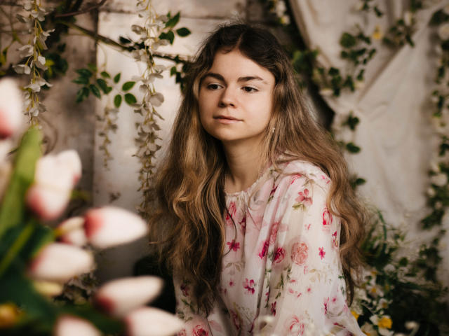 TinaRedly with long, wavy brown hair wearing a floral dress, standing amidst flowers and greenery, looking calm and confident, with white and pink tulips in the foreground.