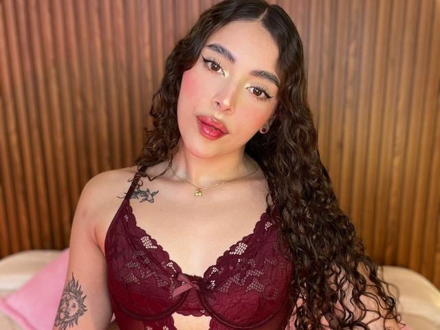 TianaVera with curly brown hair, wearing a deep red lace bra, showing her tits, red lipstick, tattoo on left arm, wooden background, looking at camera, slight smile.