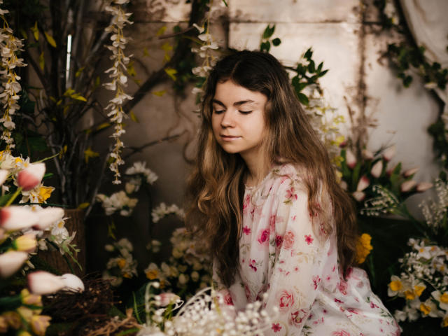 TinaRedly with long, wavy brown hair, eyes closed, wearing a floral dress, surrounded by flowers, has a serene, beautiful face, and a cute ass. Soft, natural light.