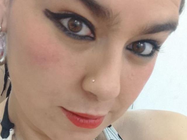 TaylorNova heeft bruine ogen, zwarte eyeliner, rode lippenstift, een neuspiercing en lichtroze wangen. Haar gezicht is in close-up te zien, met een lichte glimlach en zichtbare oren..