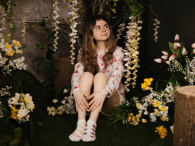 TinaRedly with wavy brown hair, floral shirt, and white socks sits among yellow and white flowers, her knees up, hands on her knees, looking off to the side.