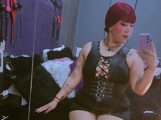 IsisStuart with red hair, black lace-up dress, showing her big tits and thick ass, tattoos, black heels, sitting in front of a mirror, taking a selfie.
