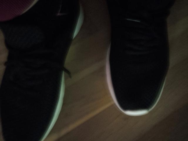 Beastbitch-hot's black sneakers with glowing white soles, laces tied, on wooden floor, slightly blurry, showing the lower part of her legs and a hint of pink pants.