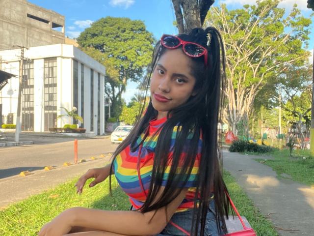 SiennaTaylor with long black hair in a high ponytail, wearing red sunglasses on her head, a rainbow-striped shirt, and a pink bag, sitting on a grassy sidewalk.