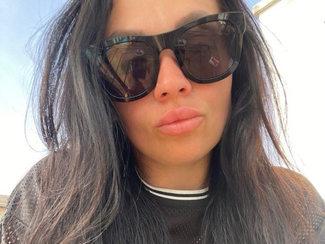 SophiaPush with long black hair, wearing black tortoiseshell sunglasses, full pink lips, and a black mesh top. Her nose is centered, and she's making a pouty face.