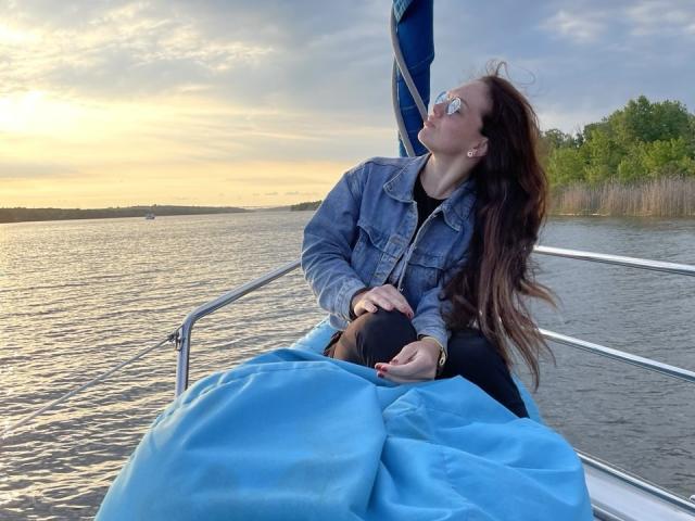 SophiaPush with long brown hair, wearing sunglasses, denim jacket, and black pants, sits on a boat, wrapped in a blue blanket, looking at the sunset, hands on lap.