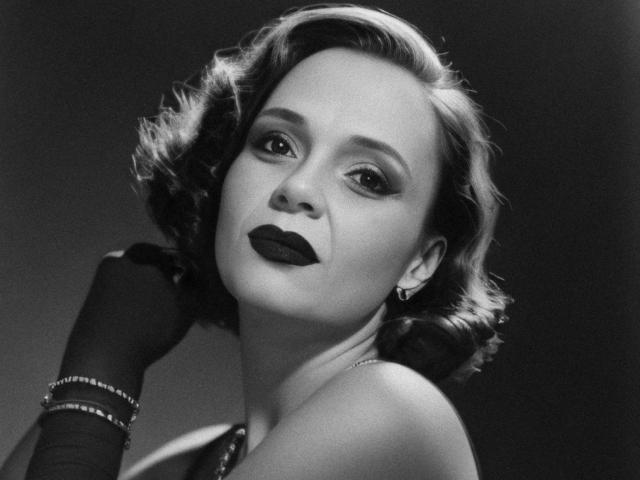 Fotografia in bianco e nero di SweeteestJulie con capelli scuri e ricci, rossetto scuro, che indossa una maglietta senza maniche, guanti e orecchini, con lo sguardo rivolto verso l'alto e la testa leggermente inclinata. Il seno è visibile, con un aspetto sensuale..