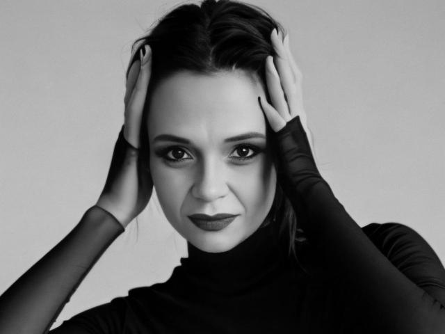SweeteestJulie has hands on head, looking serious. She's wearing a black turtleneck with sheer sleeves, black lipstick, and has dark, slicked-back hair. Black and white image.