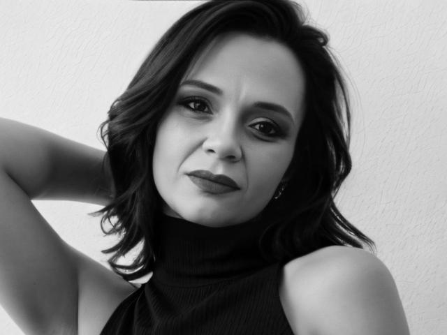 Photo en noir et blanc de SweeteestJulie, avec des cheveux noirs ondulés, portant un col roulé sans manches, les bras levés, une expression légèrement boudeuse, du rouge à lèvres foncé et de petites boucles d'oreilles..