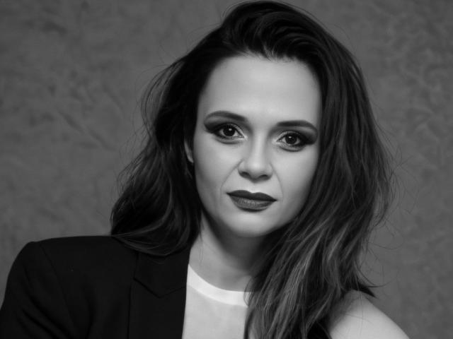 Photo en noir et blanc de SweeteestJulie, avec des cheveux noirs ondulés, un trait d'eye-liner audacieux et un rouge à lèvres foncé, portant un blazer noir par-dessus un haut blanc, regardant directement l'appareil photo..