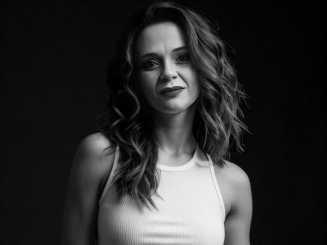 Black-and-white photo of SweeteestJulie with wavy, shoulder-length hair, wearing a white ribbed tank top, showing a subtle smile, and a slight frown on her face.
