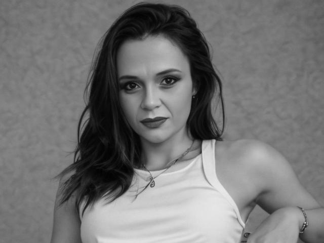 Black-and-white photo of SweeteestJulie with wavy dark hair, wearing a white tank top, showing her bare shoulders and small tits, looking slightly to the side with a neutral expression.