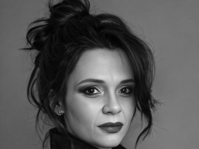 Black-and-white photo of SweeteestJulie with messy dark hair, smoky eye makeup, dark lipstick, and small pearl earrings, looking slightly to the side with a subtle smile.