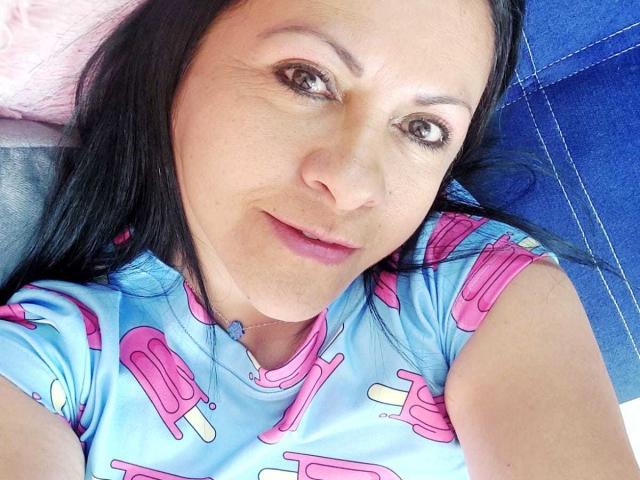 KatieHotX with pale skin, black hair, brown eyes, light pink lipstick, wearing a blue shirt with pink popsicle patterns, lying on a blue seat, subtle smile.