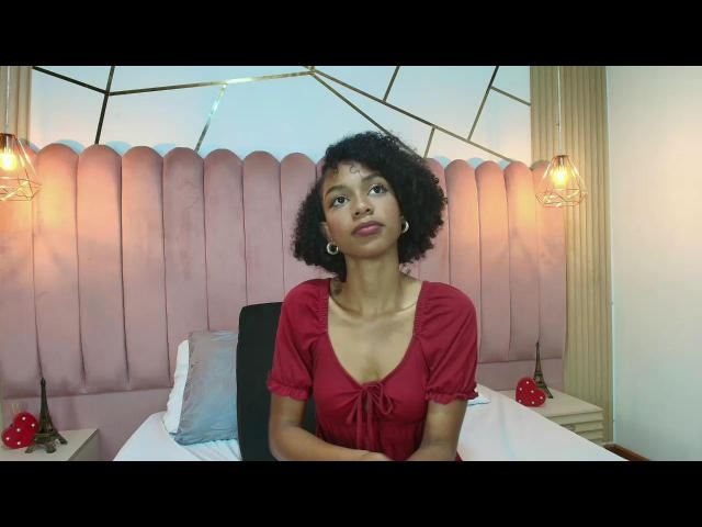 AylaCallowey with curly black hair, red dress, and hoop earrings, sitting on a bed with a pink upholstered headboard, looking slightly up and to the side, showing her tits.