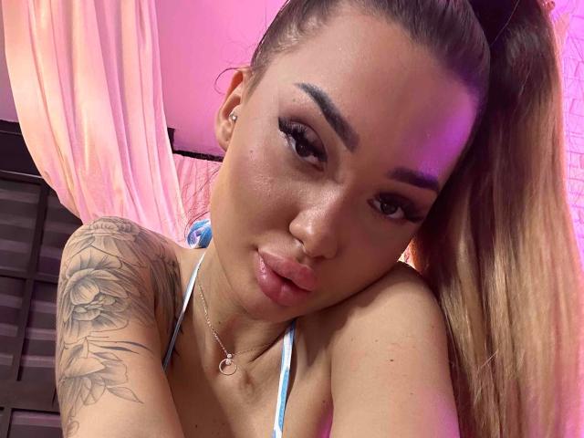 SashaFerrera with pouty lips, looking at camera, has brown hair in ponytail, white-blue bikini top, tattooed arm, and nose ring, under pink lighting. Her tits are visible.