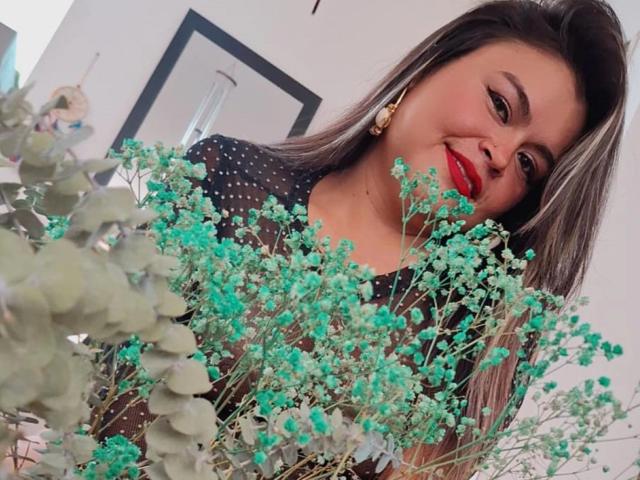 CaandyFoox with red lips, wearing a black polka-dot top, smiling while holding green baby's breath and eucalyptus flowers. She has long brown hair and gold earrings.