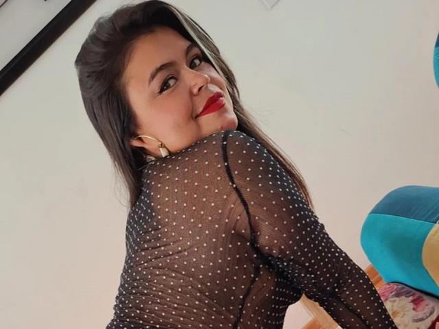 CaandyFoox with long brown hair, red lipstick, and a sheer black polka-dot top, showing her back and smiling at the camera, with gold earrings and a cute butt.