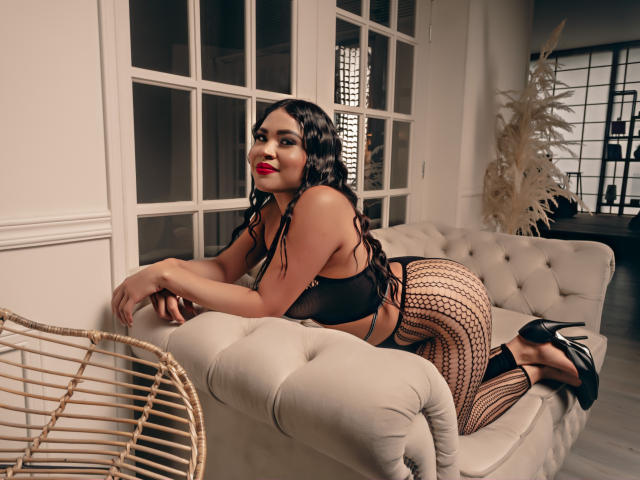 OliviGrace, com seus longos cabelos pretos e cacheados, batom vermelho, lingerie preta, calças de rede e saltos altos, ajoelhada em um sofá bege com textura, exibindo seu grande traseiro e pequenos seios..
