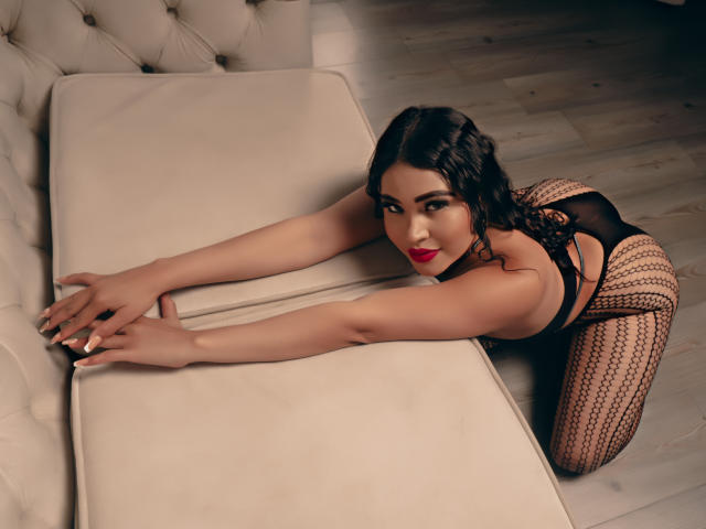 OliviGrace de quatro, vestindo lingerie preta com meias estampadas, braços estendidos sobre um travesseiro bege, olhando para cima com batom vermelho e cabelo escuro ondulado..