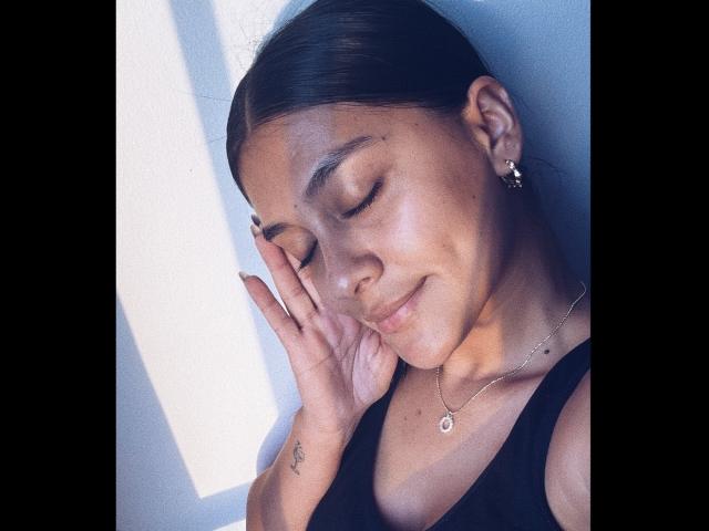 ThaliaAres with closed eyes, smiling slightly, wearing a black top, hand touching face, small tattoo on wrist, earrings, necklace, and brown hair pulled back. Soft sunlight on her face.