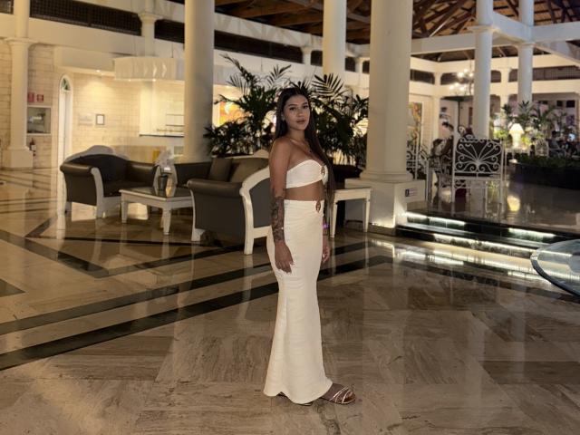 ThaliaAres stands in a fancy hotel lobby, wearing a white strapless top and white, form-fitting, long dress, showing off her ass and tattoos, with brown sandals on.