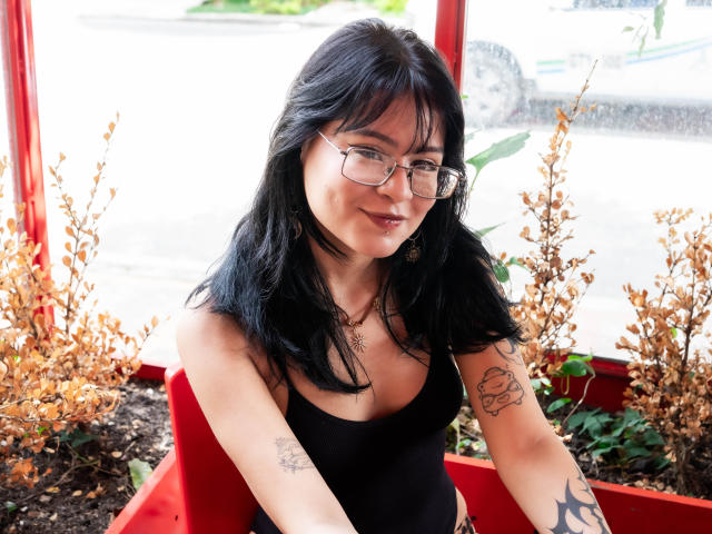 ElizabethRoux with black hair, glasses, and a black tank top, showing tattoos on her arms, sitting in a red chair, smiling, with dry plants in the background.