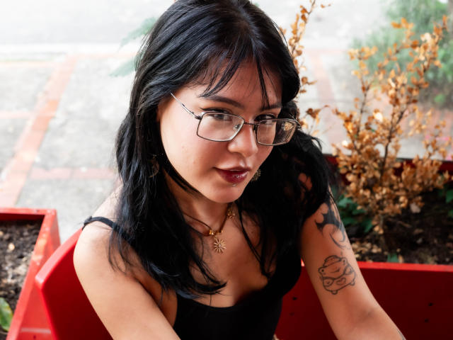 ElizabethRoux with black hair, glasses, and a black top, showing a tattoo on her right arm, wearing a sun necklace, looking up with a slight smile.
