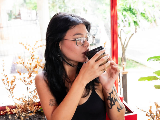 Elizabeth Roux, con i capelli neri e gli occhiali, sorseggia da una tazza di caffè nero, mostrando un tatuaggio sul braccio destro, indossa una canotta nera, in interni con piante..