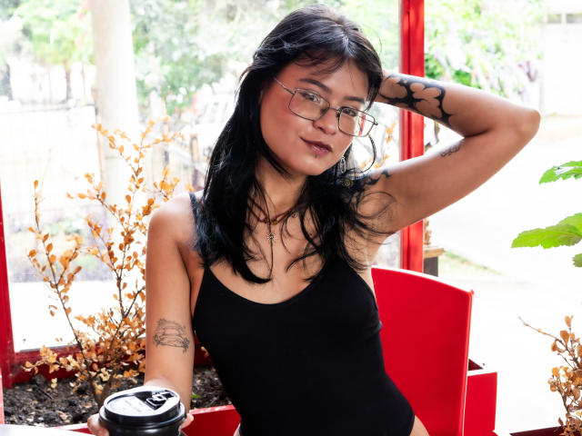 Elizabeth Roux, con capelli neri, occhiali e tatuaggi, indossa una canotta nera, tiene in mano una tazza di caffè, con un braccio alzato, sorridendo, in un ambiente rosso e verde..