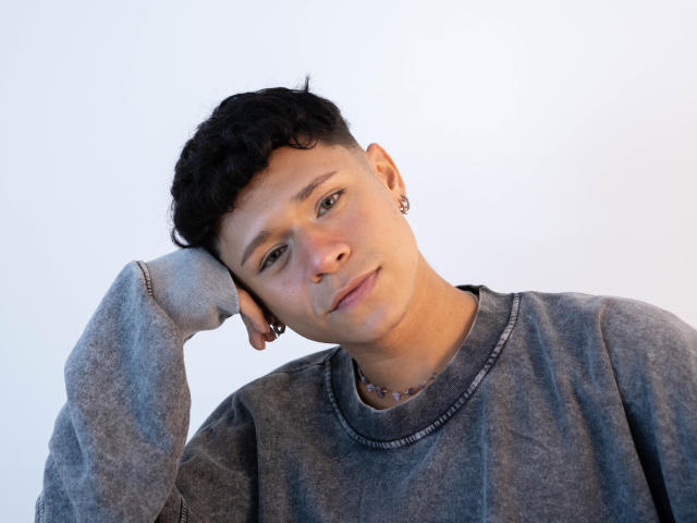 DanteEvans with short curly black hair, wearing a grey sweatshirt, resting his head on his hand, looking slightly to the side, earrings visible, soft natural light.