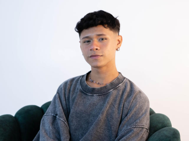 DanteEvans with short black curly hair, wearing a gray sweatshirt, small hoop earring, and necklace, sitting on a green chair, looking slightly to the side.
