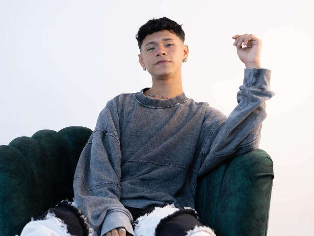 DanteEvans with short black hair, wearing a gray sweatshirt and black ripped jeans, sitting on a green velvet chair, raising his right arm, showing a tattoo on his neck.