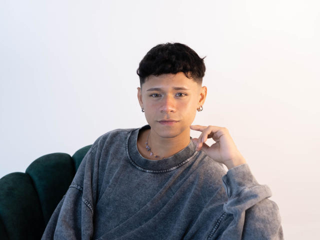 DanteEvans with short black curly hair, wearing a grey sweater, small earrings, necklace, green velvet chair, hand near chin, white background, looking at camera with a neutral expression.