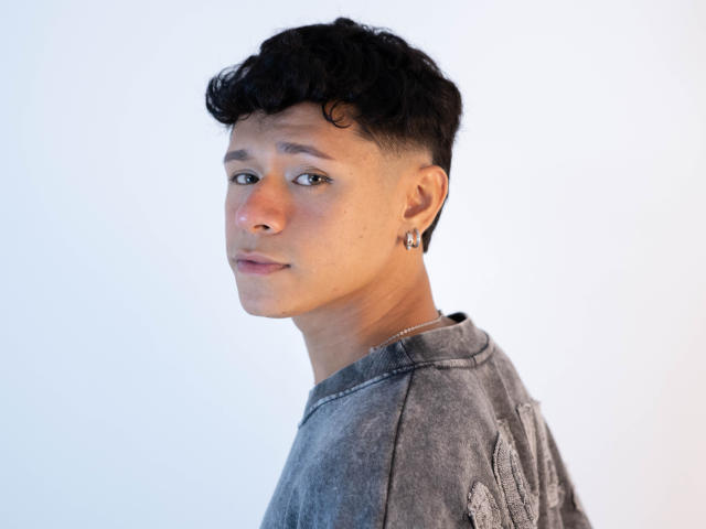 DanteEvans, short curly black hair, light skin, small nose ring, ear gauge, grey textured shirt, looking over shoulder, slight smile, soft light, casual style.