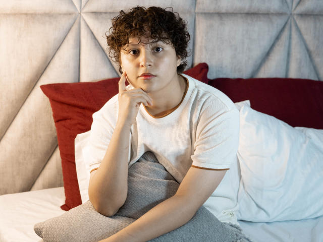 MadisonTaylorr, met krullend bruin haar, een wit T-shirt en een serieuze uitdrukking, ligt op bed, met haar hand onder haar kin. Er ligt een grijs kussen, rode kussens en witte beddengoed. Haar achterkant is gedeeltelijk zichtbaar..