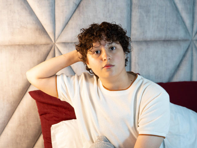 MadisonTaylorr, avec ses cheveux bruns bouclés, son t-shirt blanc et sa main dans les cheveux, a l'air fatiguée et ennuyée, assise sur un lit avec un oreiller rouge et une tête de lit grise..