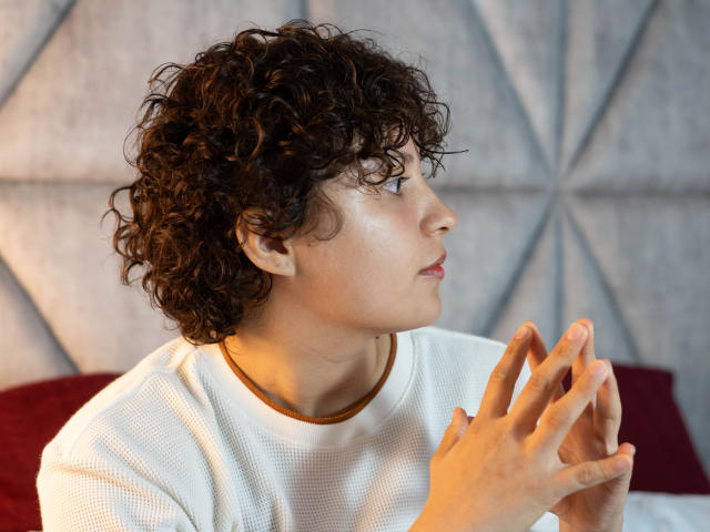 MadisonTaylorr, avec ses cheveux bruns bouclés, son chemisier blanc texturé et son col marron, les mains jointes, regardant sur le côté avec une expression légèrement pensive, une lumière douce sur son visage..
