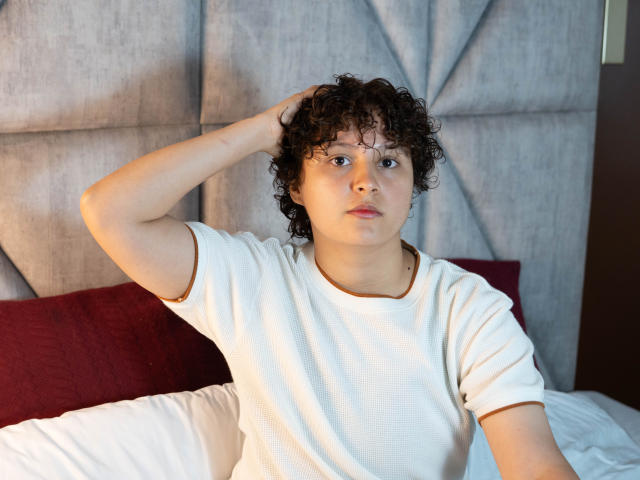 MadisonTaylorr heeft krullend bruin haar, een wit T-shirt, en heeft haar hand in haar haar, terwijl ze op een bed zit met een rood kussen en witte lakens. Haar gezicht heeft een neutrale uitdrukking..